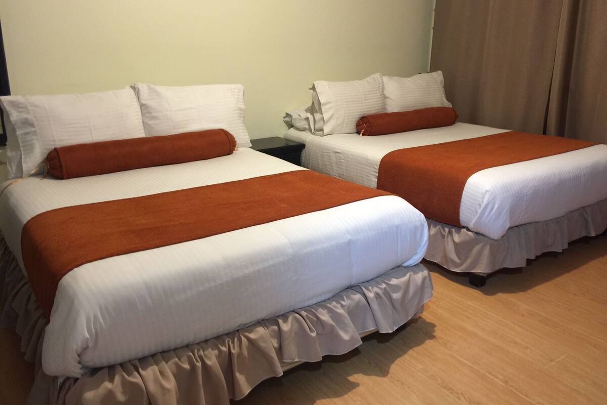 Two twin beds with white linens and brown headboards, a wooden floor, and a plain wall.