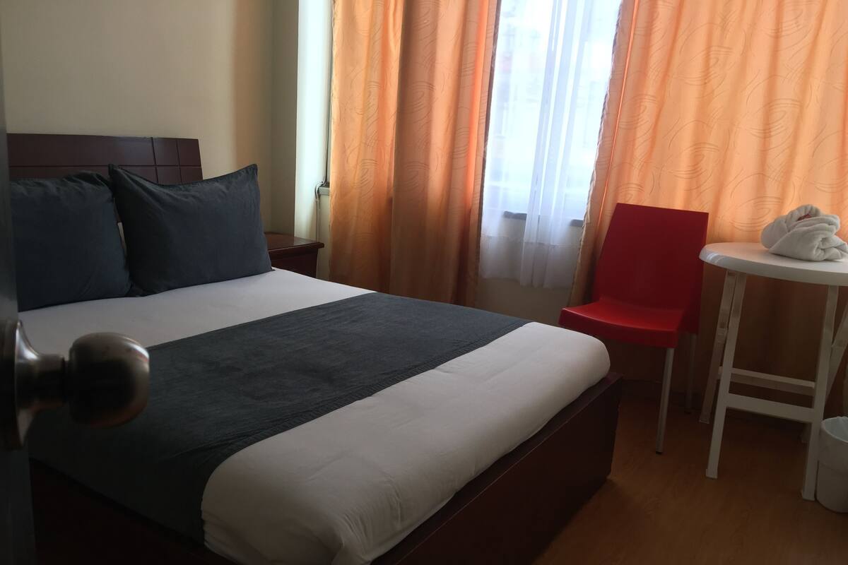A hotel room with a bed, a red chair, a small table with a towel, and curtains.