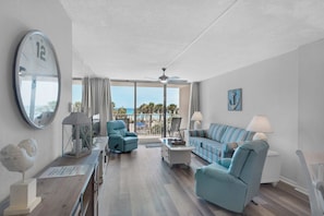A spacious living room with a large clock, a sofa, a chair, and a view of the ocean. - Dunes Of Panama B208 (Panama City Beach)