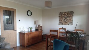 Un salon comprenant un meuble en bois, un fauteuil bleu, une table à manger avec des chaises, une horloge au mur et un élément décoratif mural.