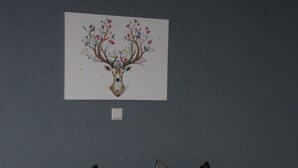 Een tweepersoonsbed met een geruit beddegoed en passende kussens, een nachtkastje met een glas, en een aan de muur hangend beeld van een hert met hoorns en bloemen.
