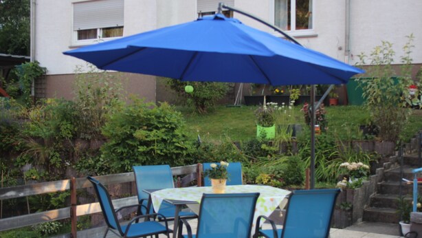 Een terras buiten met een tafel en stoelen, een blauwe parasol, en een tuin op de achtergrond.