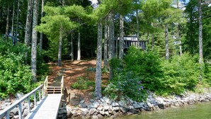 A wooden dock leading to a house surrounded by tall trees and a rocky shoreline.