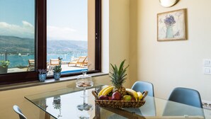 Een eethoek met een glazen tafel, een fruitmand en uitzicht op zee door een groot raam.