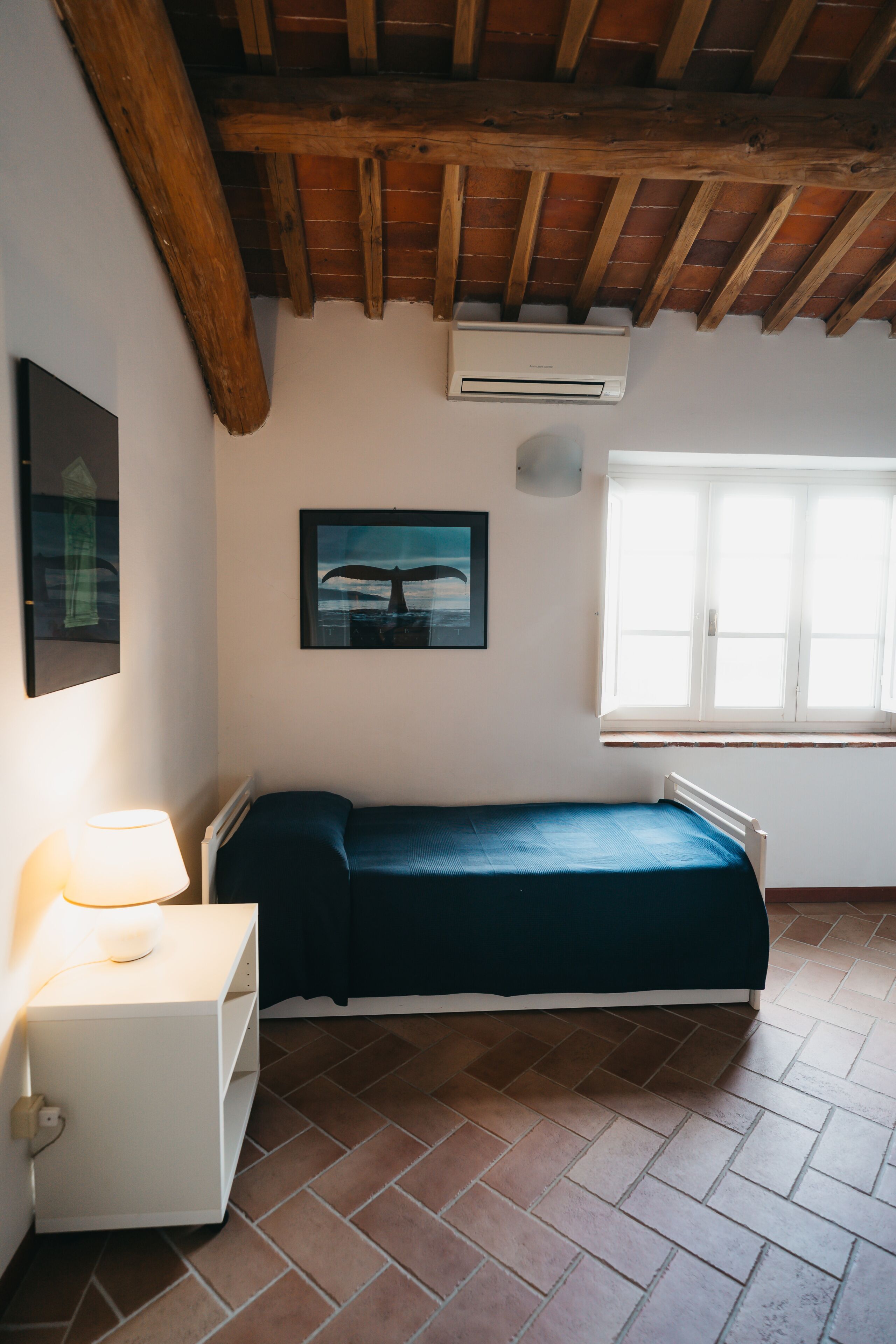Une petite pièce comprenant un lit, un climatiseur fixé au mur, une fenêtre et un tableau accroché au mur.