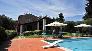 Una piscina con tumbonas y sombrillas, un edificio de piedra con techo de tejas y un jardín bien cuidado.