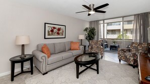 A living room with a grey sofa, two armchairs, a coffee table, a ceiling fan, a framed floral artwork, a potted plant, and a view of the outside balcony. - Home At The Maya.... (Scottsdale)