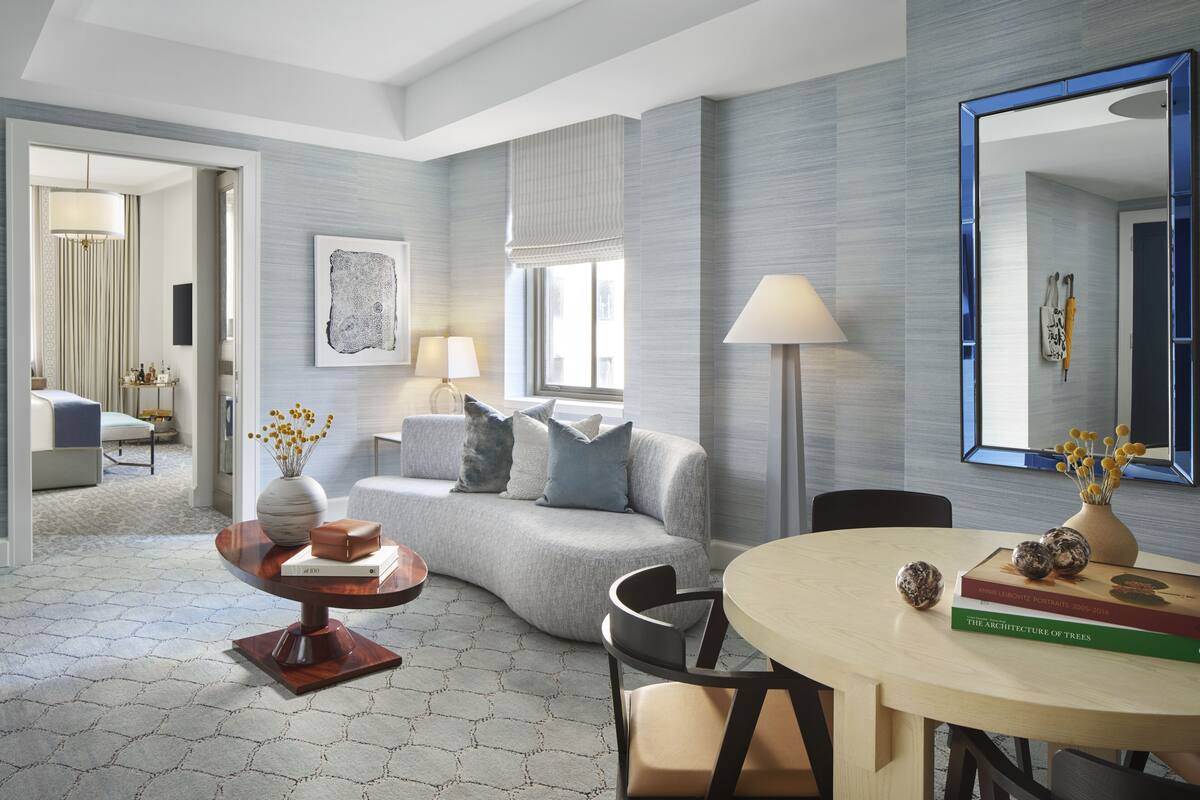 A modern living room with a grey sofa, a round coffee table, and a dining table with chairs. There is a large mirror on the wall and a window with blinds.