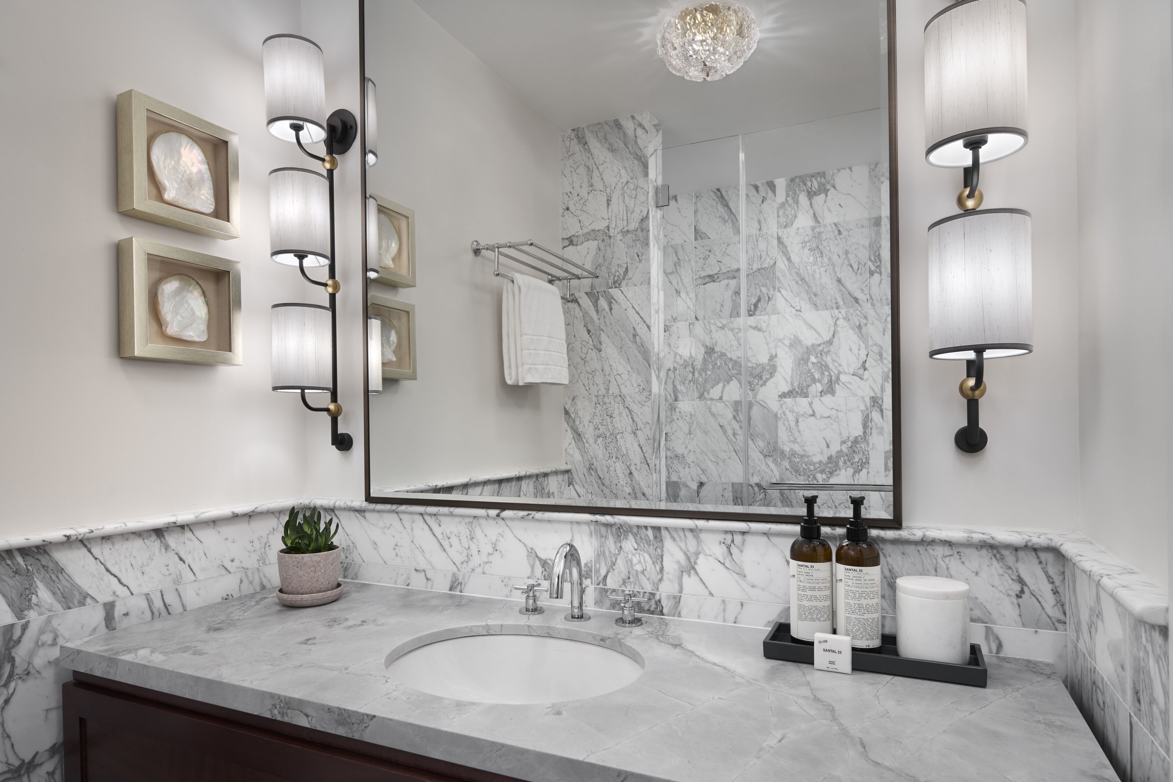 Un bagno con un lavabo in marmo, due lampade a parete, una pianta in vaso e uno specchio che riflette l'area doccia.
