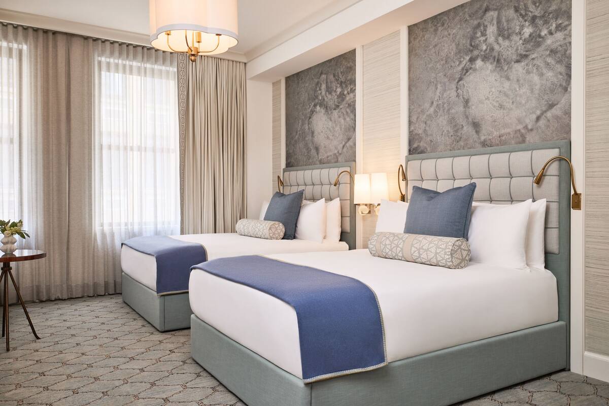 A hotel room with two beds, a chandelier, a large abstract wall painting, and a view of the outdoors through sheer curtains.