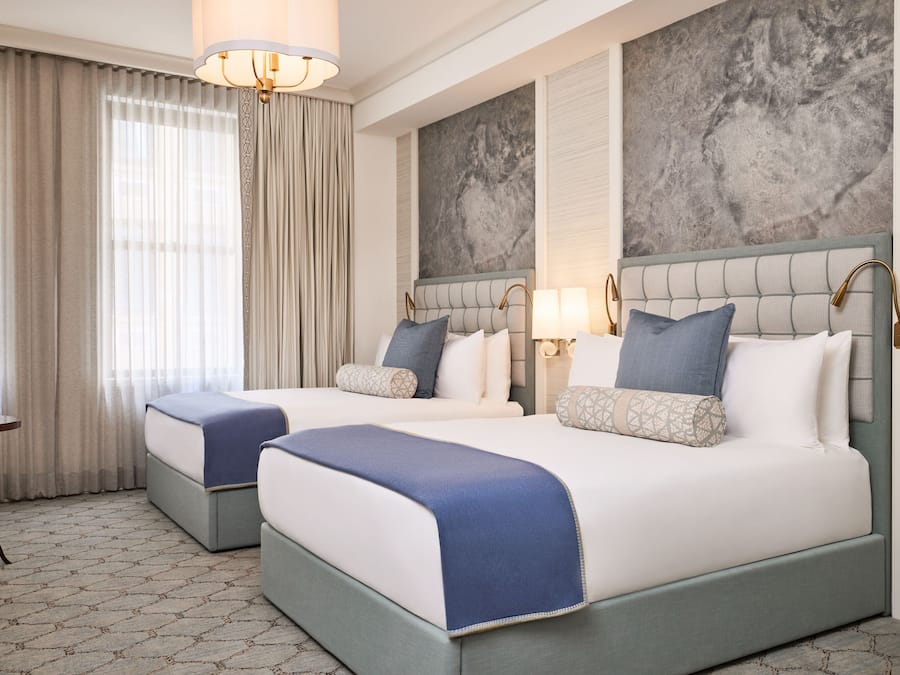 Ein Hotelzimmer mit zwei Betten, einer Kandelaber, einem großen abstrakten Wandbild und Blick ins Freie durch transparente Vorhänge.