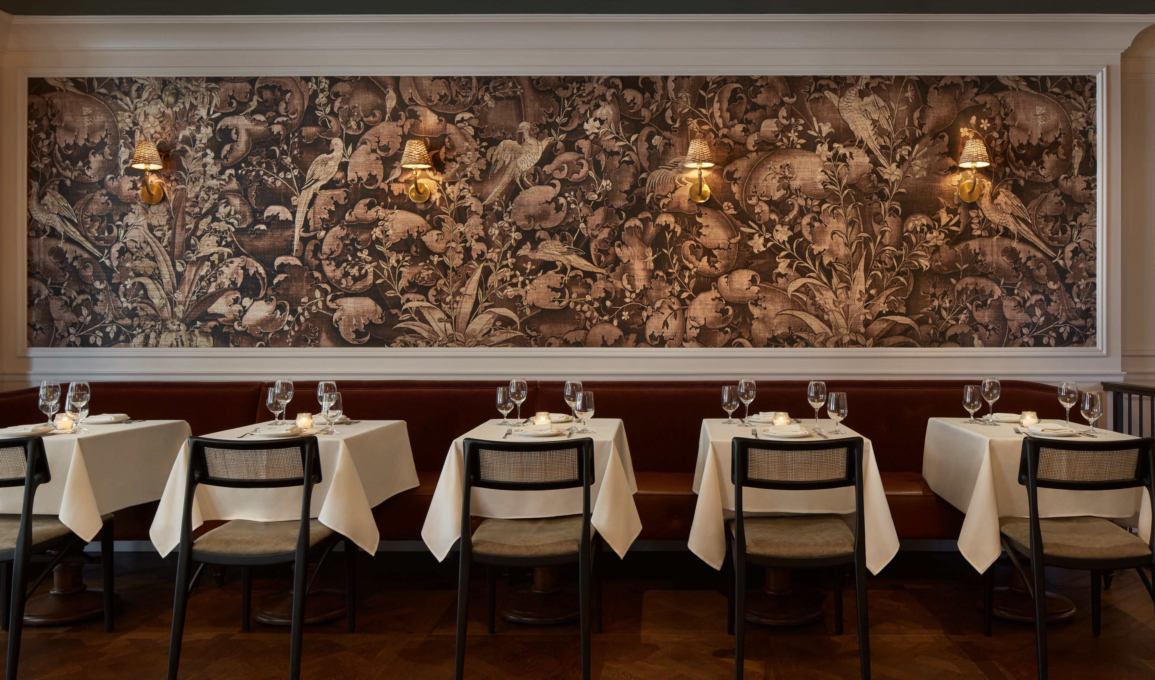 A restaurant with six tables, each with a white tablecloth and a candle. The walls are adorned with a large, detailed floral tapestry.