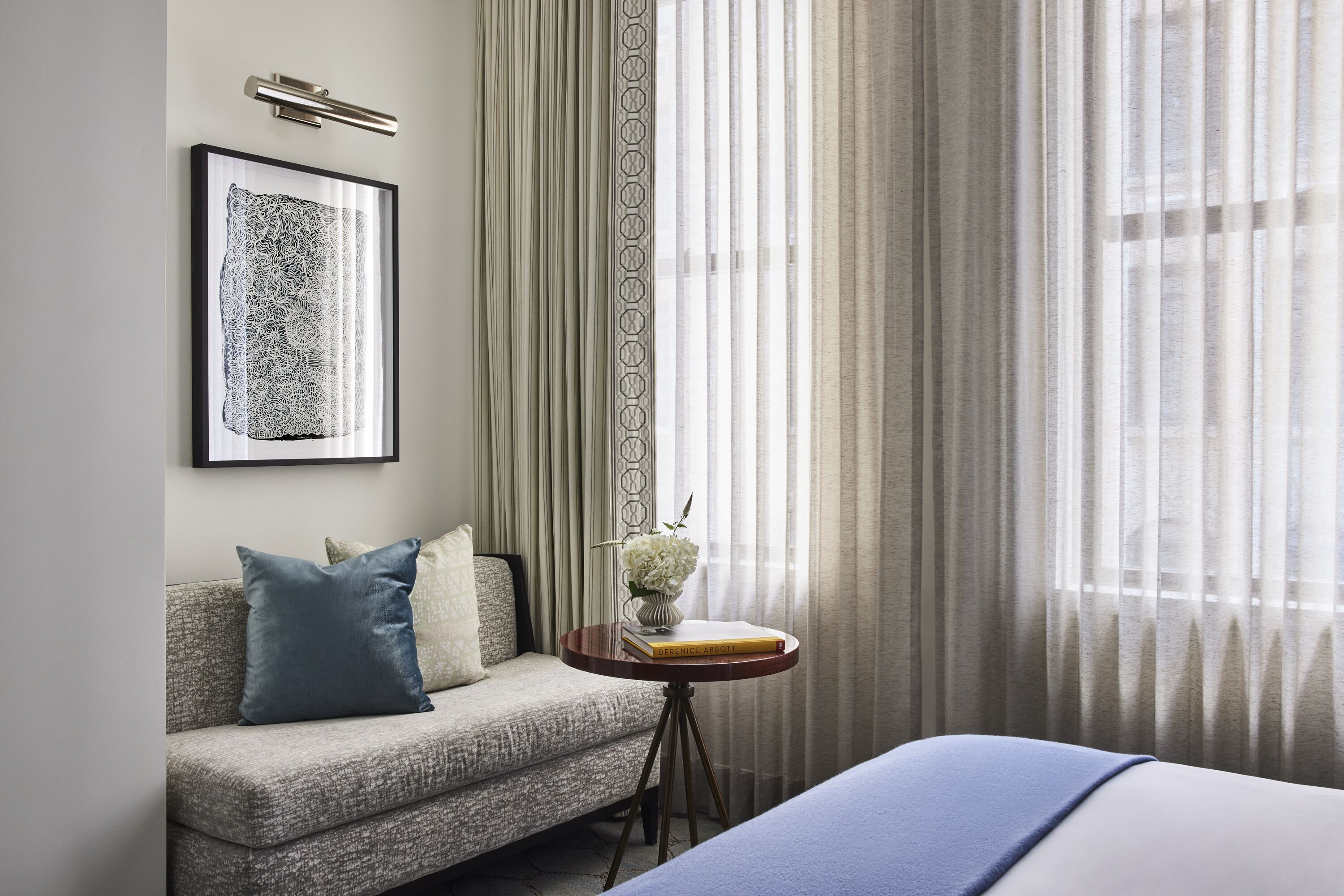 A hotel room with a sofa, a small round table with a book and a vase, and a bed with a blue blanket.