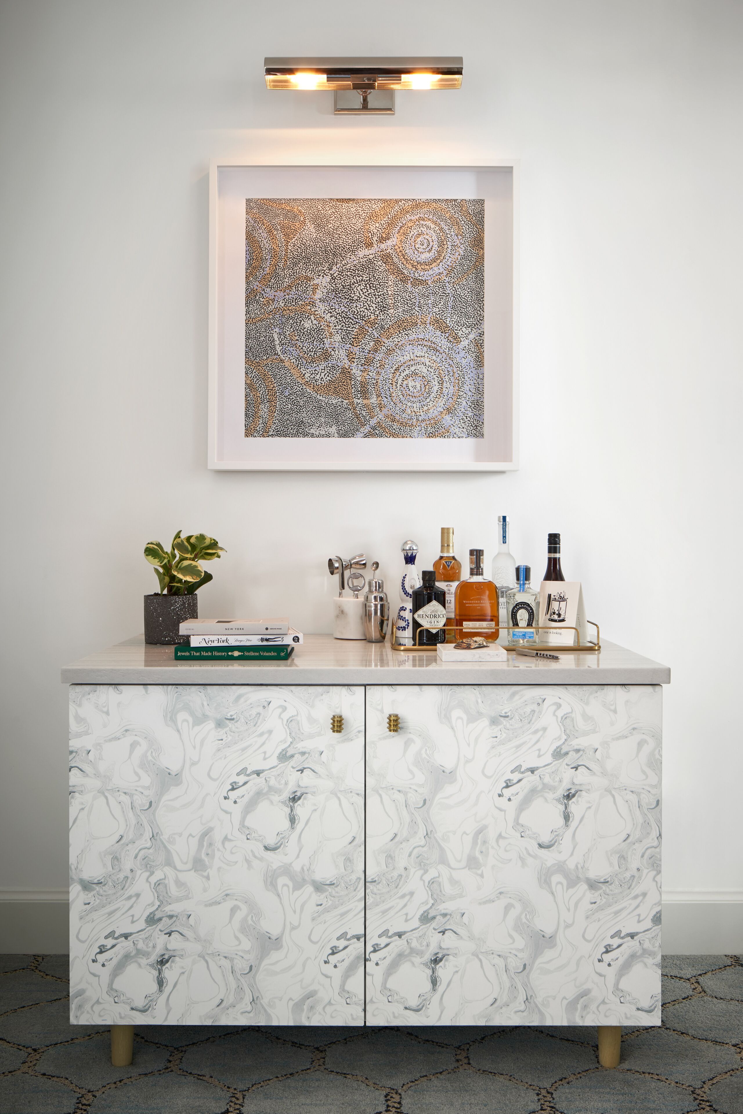 A marble-patterned cabinet with a framed abstract painting above it, a potted plant, and various bottles and books on the countertop.