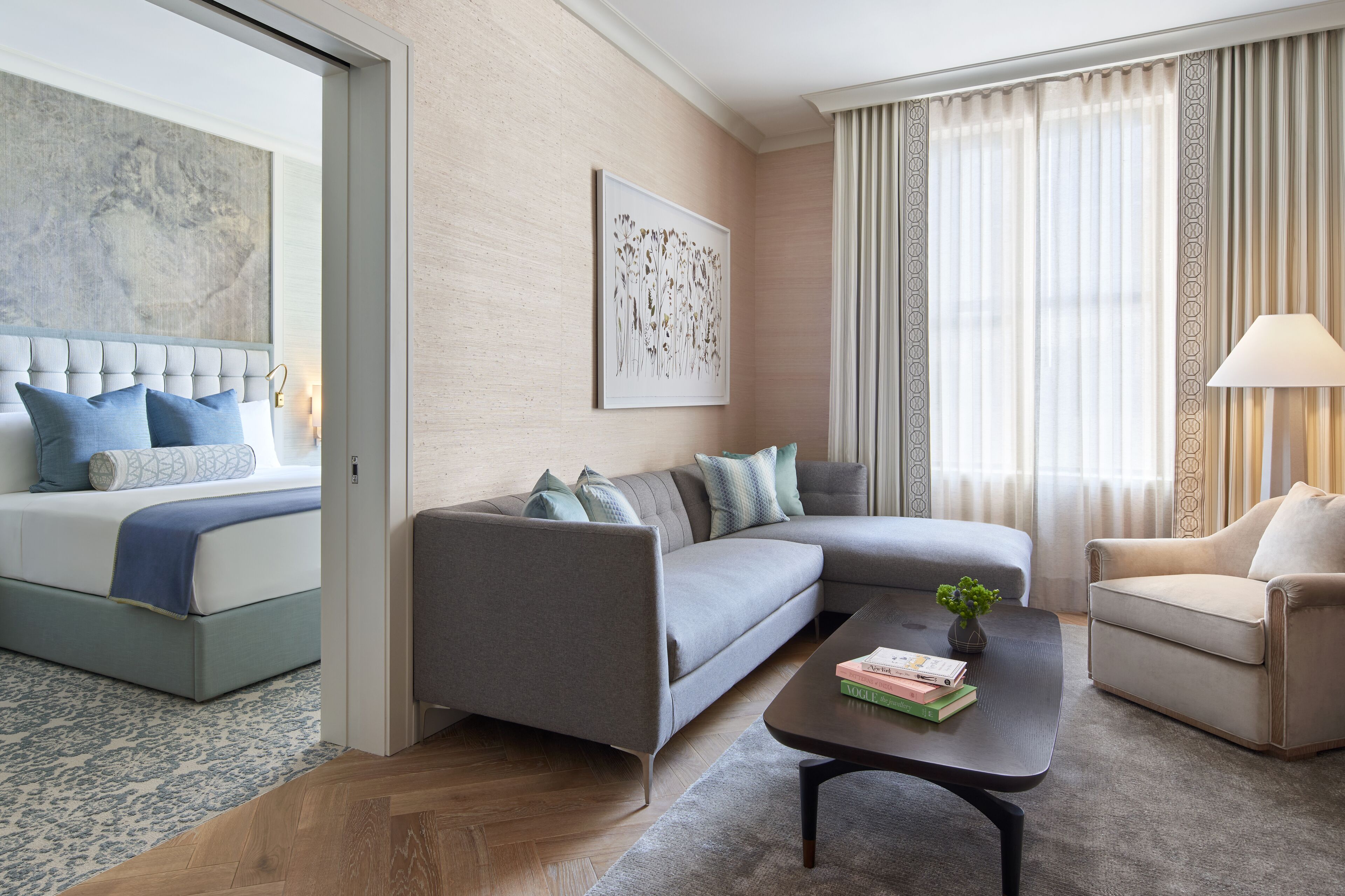 A modern hotel room with a bed, grey sofa, armchair, and a coffee table with books.
