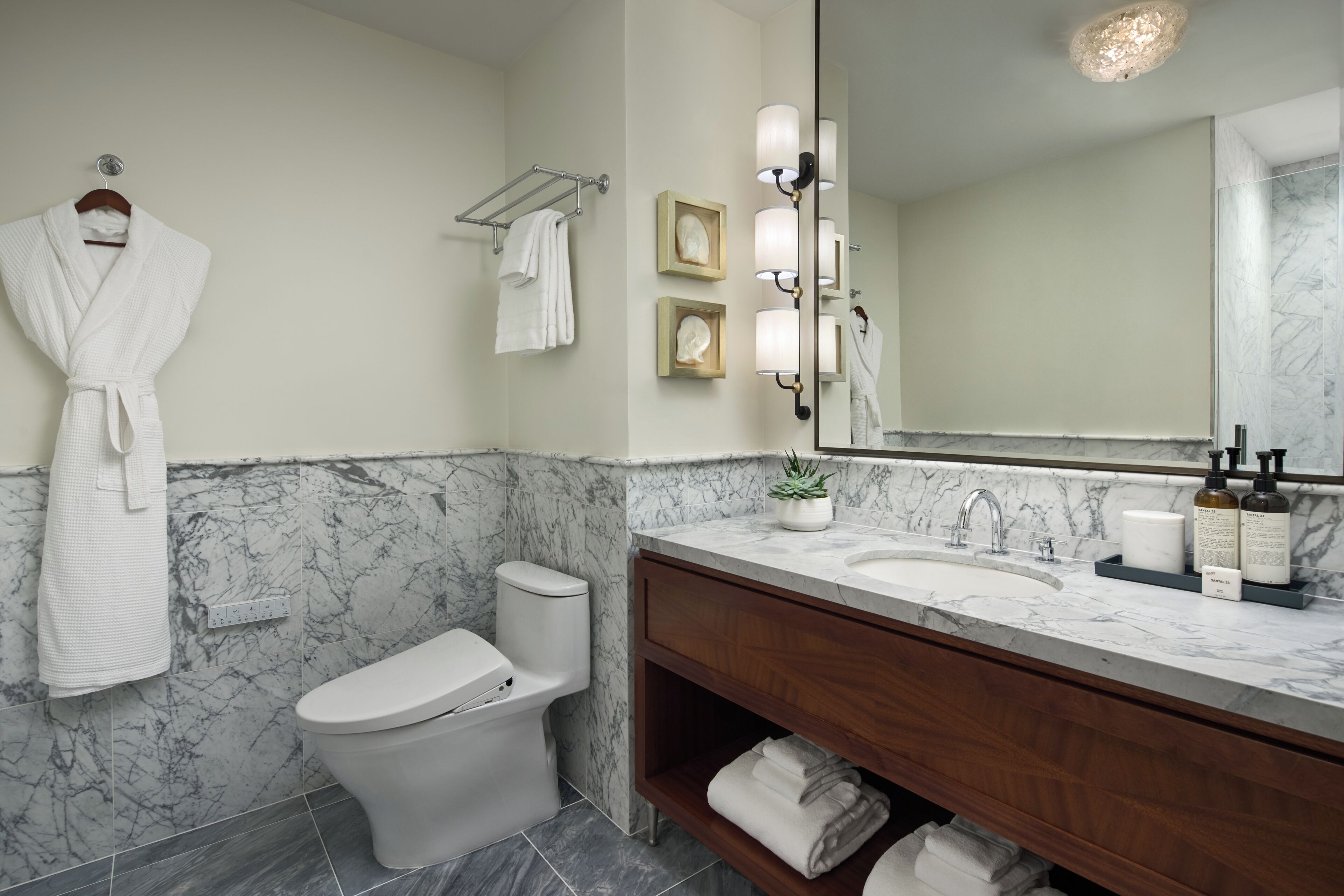 A bathroom with a marble wall, a white toilet, a vanity with a mirror, and a robe hanging on the wall.
