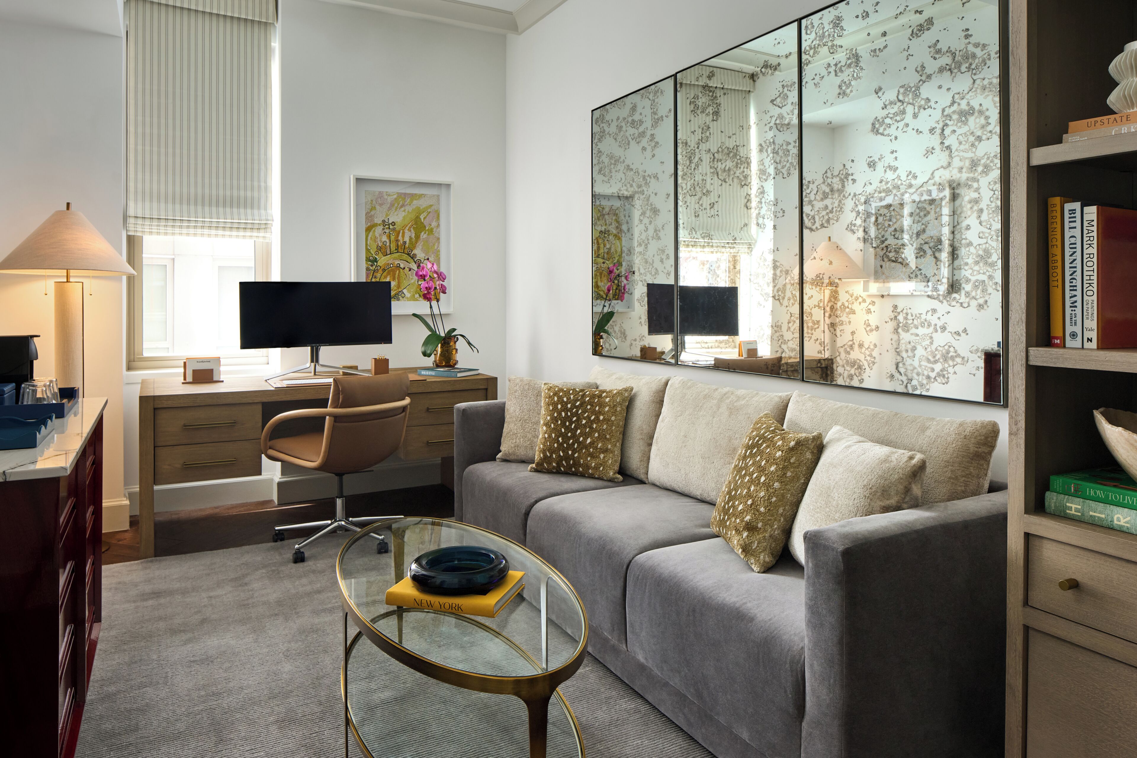 Ein modernes Wohnzimmer mit einem grauen Sofa, einem Glastisch, einem Schreibtisch mit einem Computer und einem Bücherregal.