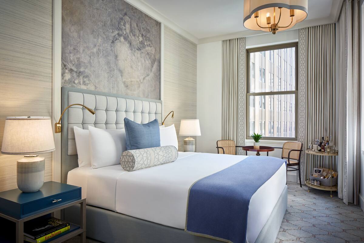 A modern hotel room with a large bed, two bedside tables, a small side table, a window with curtains, and a view of the city.
