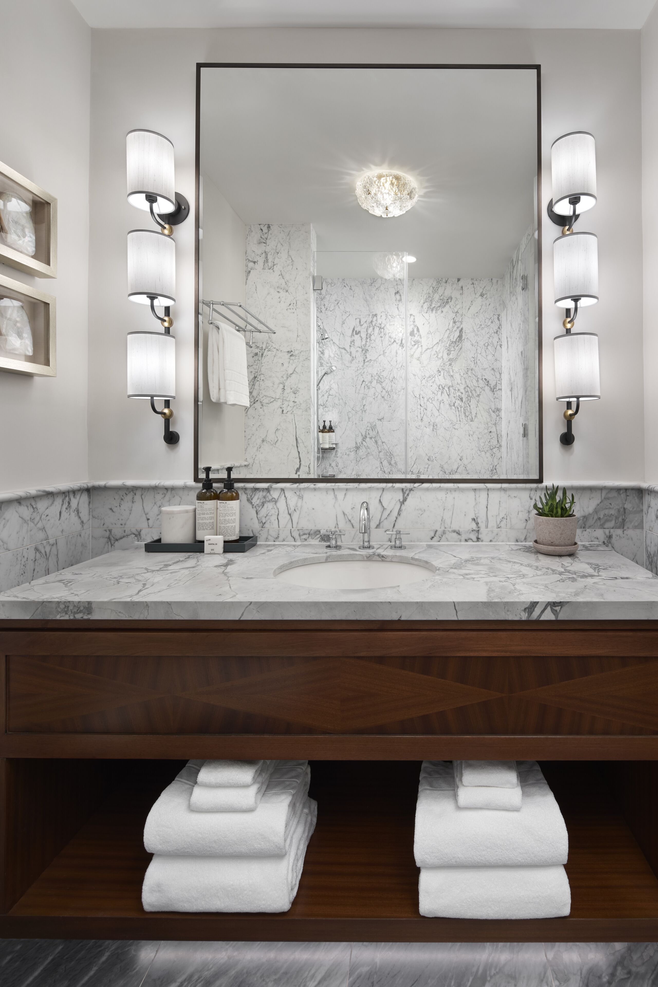 A modern bathroom with a marble countertop, a large mirror, wall-mounted sconces, and a towel rack.