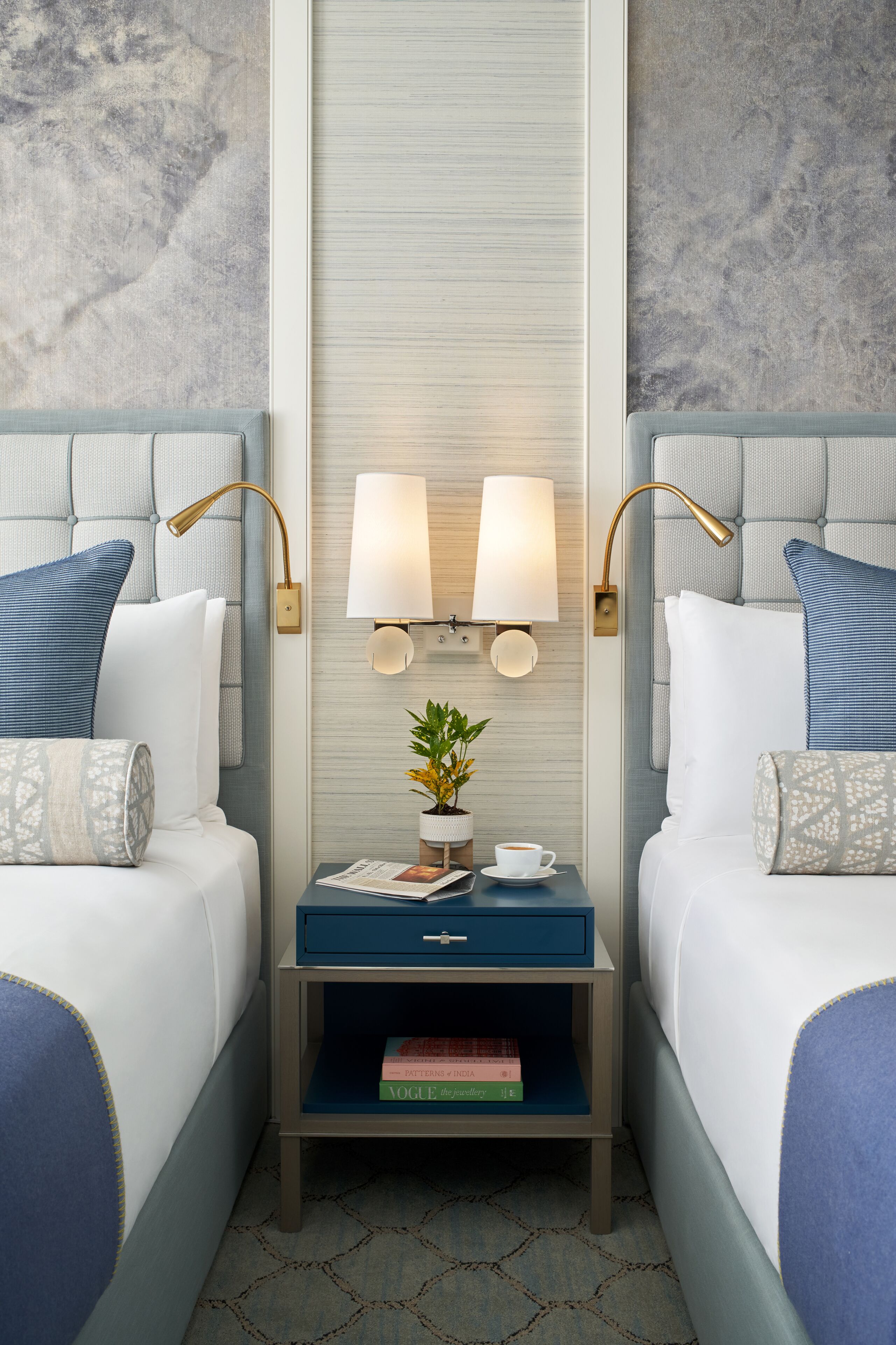 A hotel room with two beds, a nightstand with a lamp, a small table with a plant, and a wall-mounted light fixture.