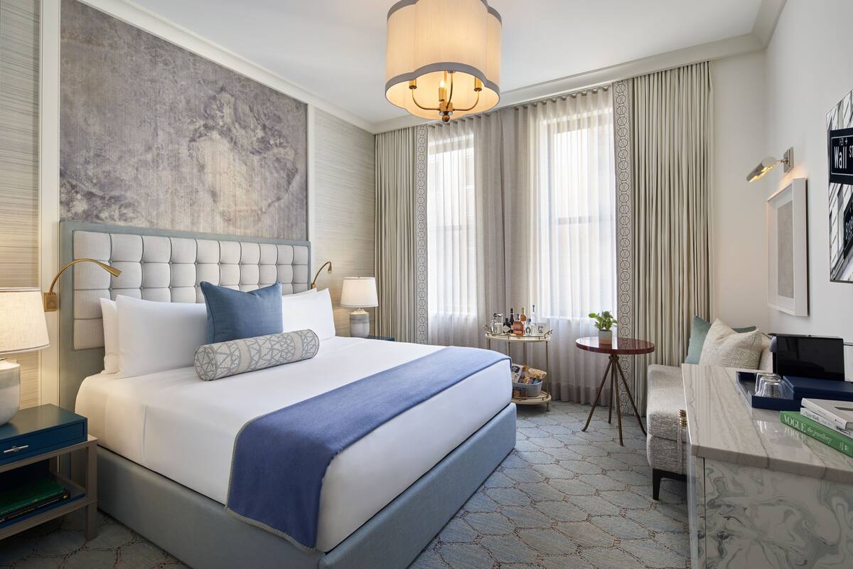 A hotel room with a large bed, a desk, a chair, a small table, and a large window with curtains.