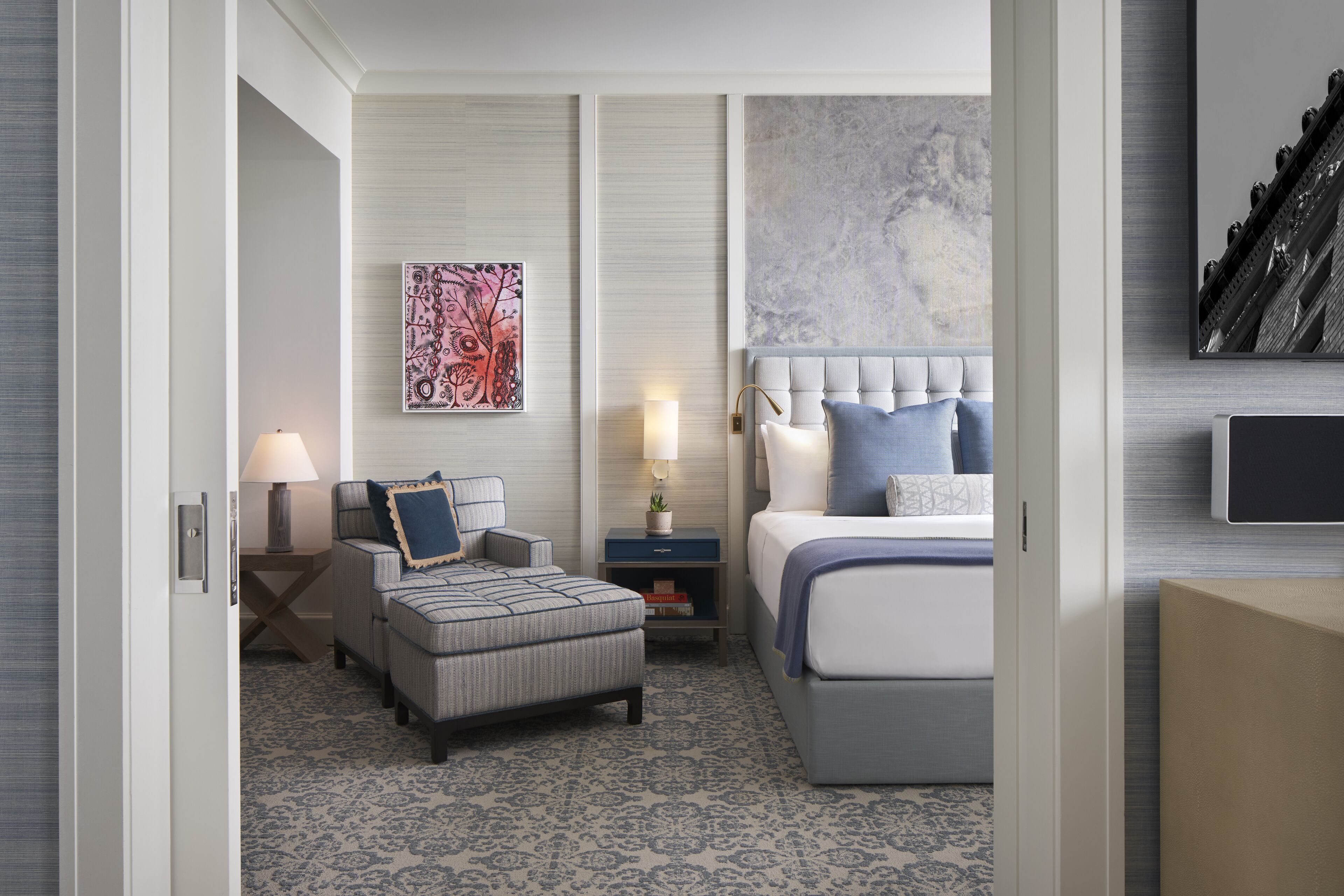 A modern hotel room with a large bed, a seating area with a sofa and armchairs, a small table with a lamp, and a wall-mounted TV.