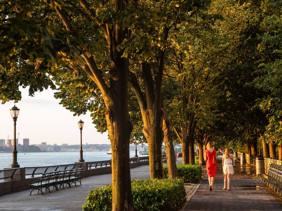 A waterfront promenade with benches and lamp posts, lined with trees and a river in the background.