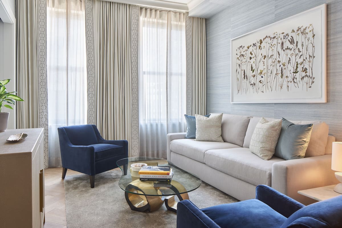 A modern living room with a blue armchair, a glass coffee table, a beige sofa, and a large framed botanical print on the wall.