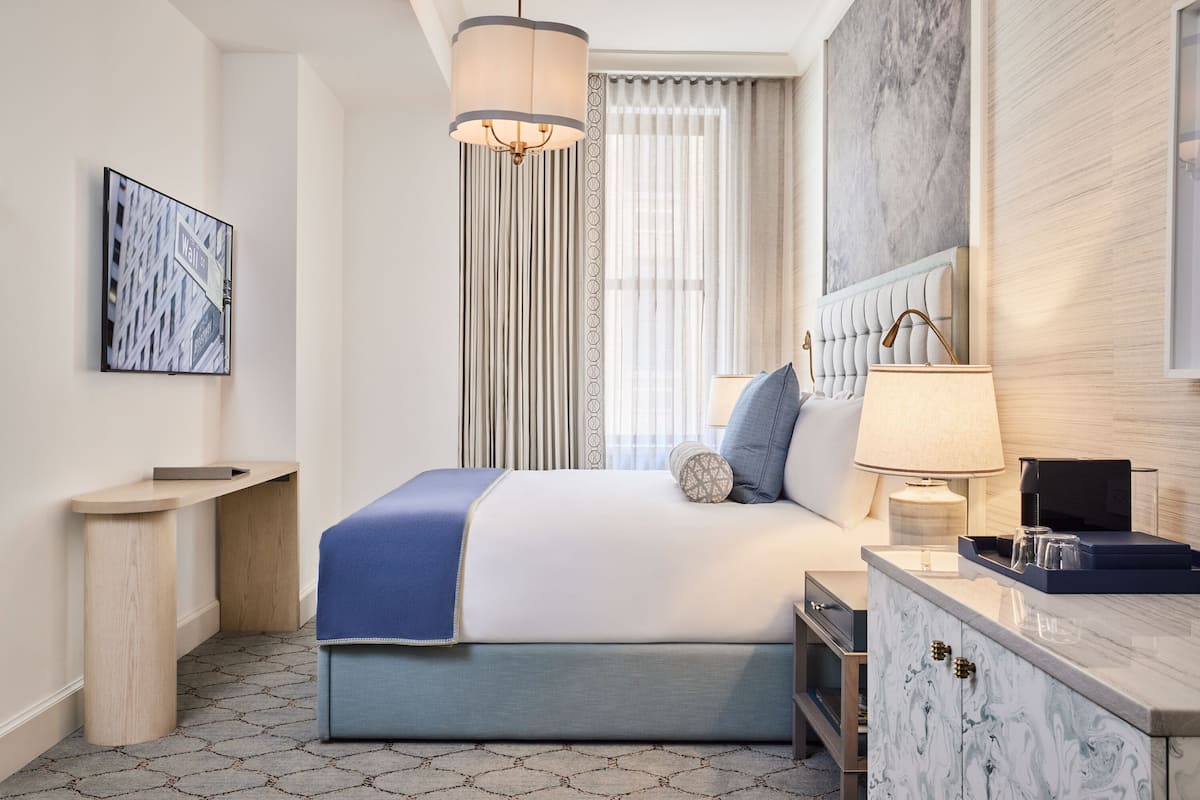 A modern hotel room with a large bed, a bedside table, a lamp, and a view of the city through the window.