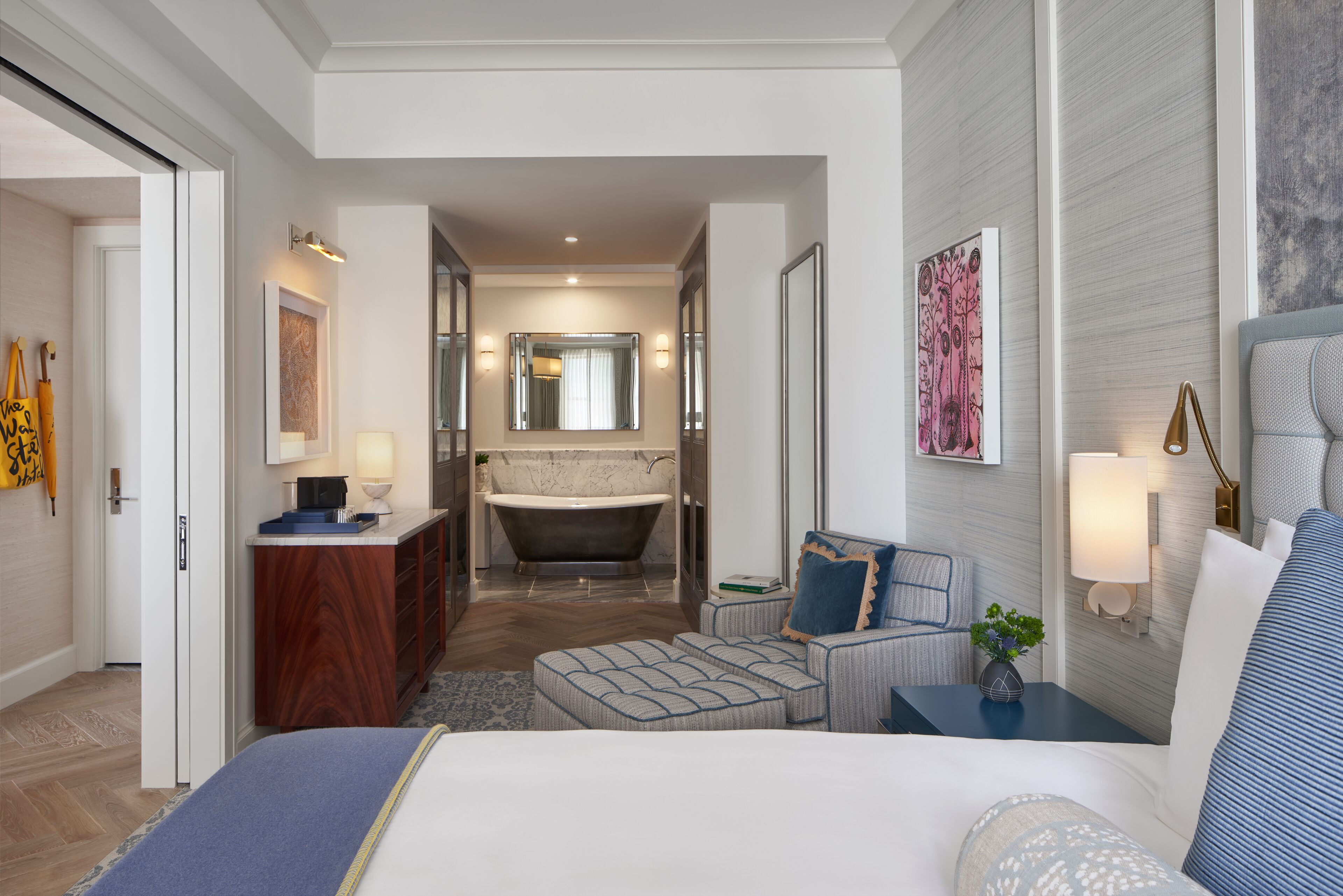 A modern hotel room with a large bed, a sitting area with a small table, a bathtub, and a separate bathroom with a vanity and mirror.