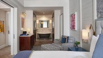 A modern hotel room with a large bed, a sitting area with a small table, a bathtub, and a separate bathroom with a vanity and mirror.