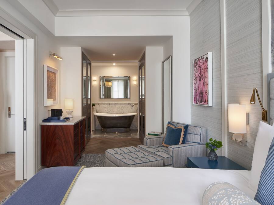A modern hotel room with a large bed, a sitting area with a small table, a bathtub, and a separate bathroom with a vanity and mirror.
