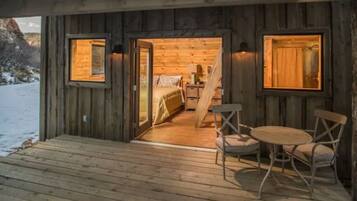 Una cabaña de madera con un porche de madera, un comedor con mesa y sillas, y un dormitorio con cama y cómoda.