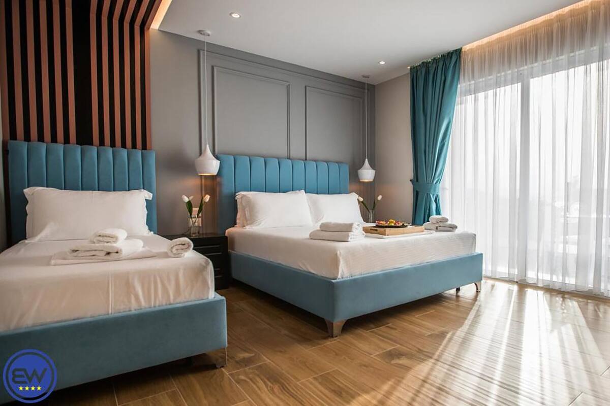 A hotel room with two beds, a wooden floor, a large window with curtains, and a wall with vertical stripes.