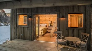 A wooden cabin with a wooden deck, a dining area with a table and chairs, and a bedroom with a bed and a dresser.