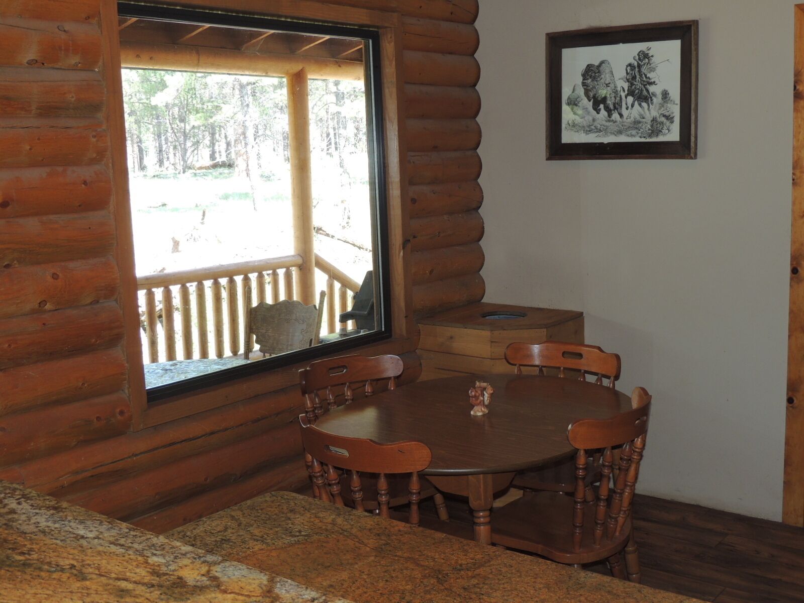 L’intérieur d’une cabane en bois, avec une table et des chaises, une fenêtre donnant sur une terrasse et un tableau encadré accroché au mur.