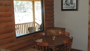 Innenansicht einer Holzhütte mit Esstisch und Stühlen, einem Fenster mit Blick auf eine Terrasse und einem gerahmten Bild an der Wand.