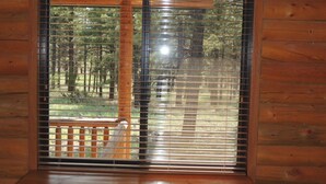Eine Holzhütte mit einem großen Fenster, ausgestattet mit horizontalen Jalousien, einer Holzdiele und Blick auf ein bewaldetes Gebiet.