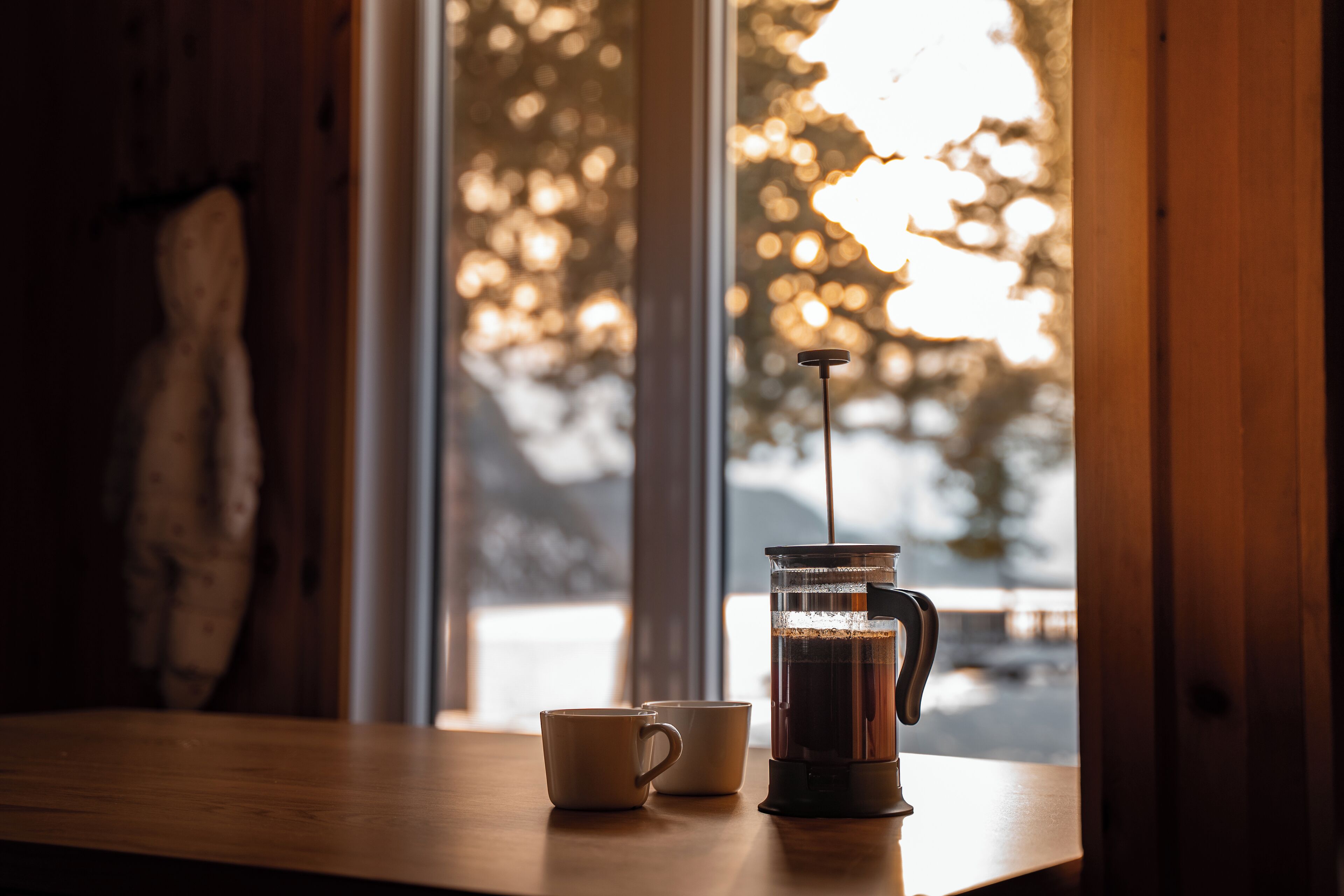 A coffee maker on a wooden table with two cups beside it, near a window with a view of trees.