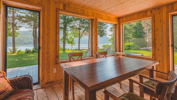 A wooden interior with a large table and chairs, large windows with a view of a lake and trees. - Le 2800 du Parc (Shawinigan)