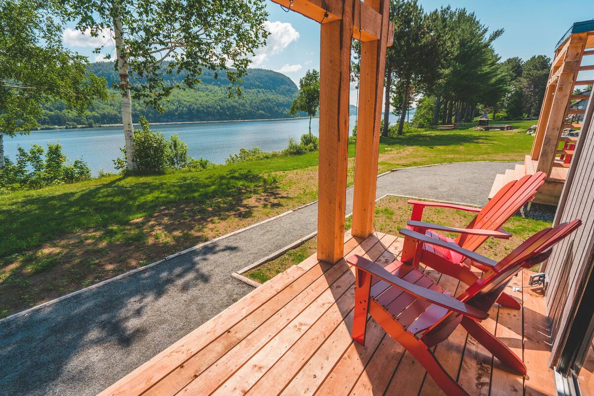 Um deck de madeira com cadeiras Adirondack vermelhas, com vista para um lago e uma colina arborizada ao fundo.