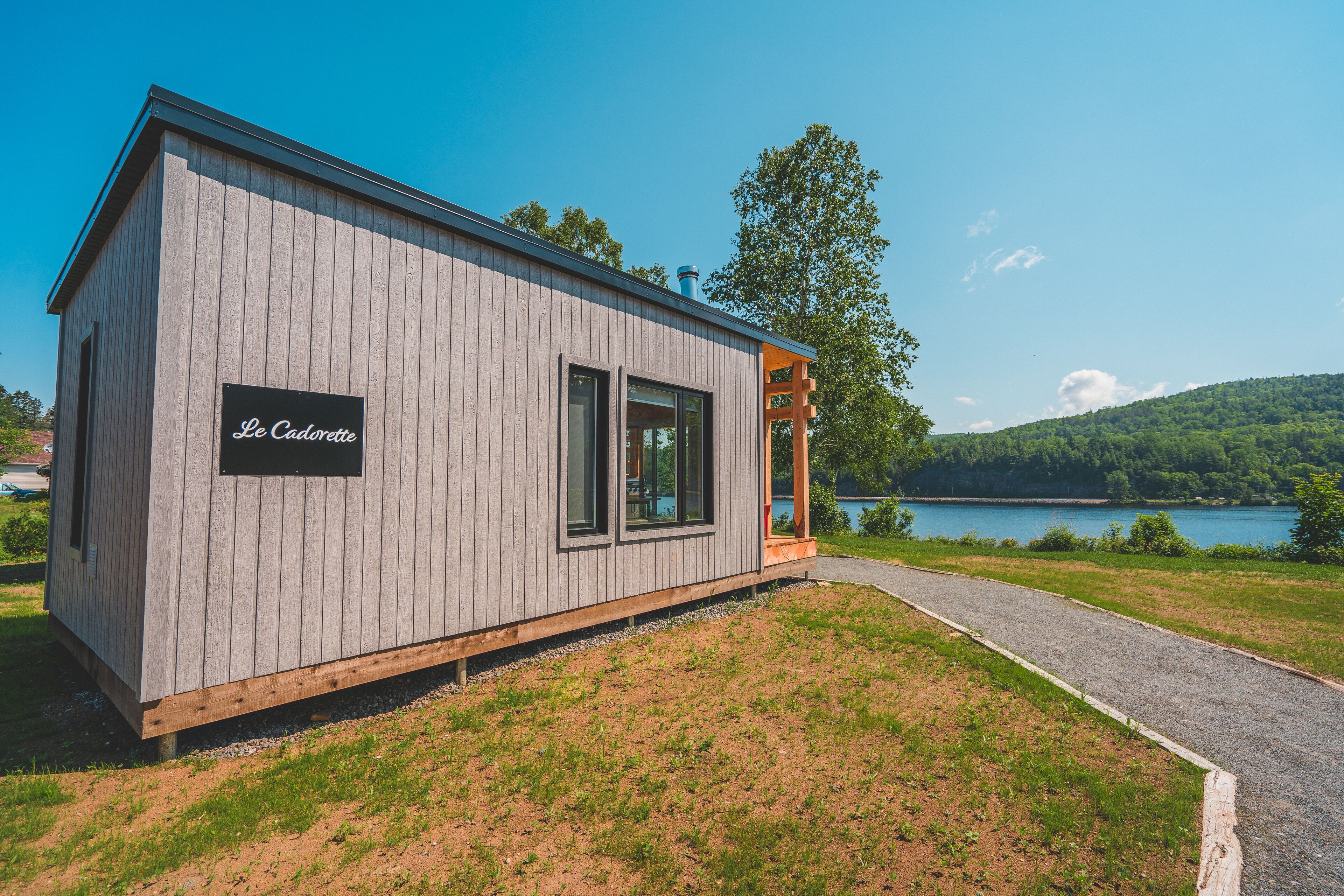 Une cabane en bois avec une enseigne indiquant « Le Cadorette », un chemin de gravier et un lac en arrière-plan.