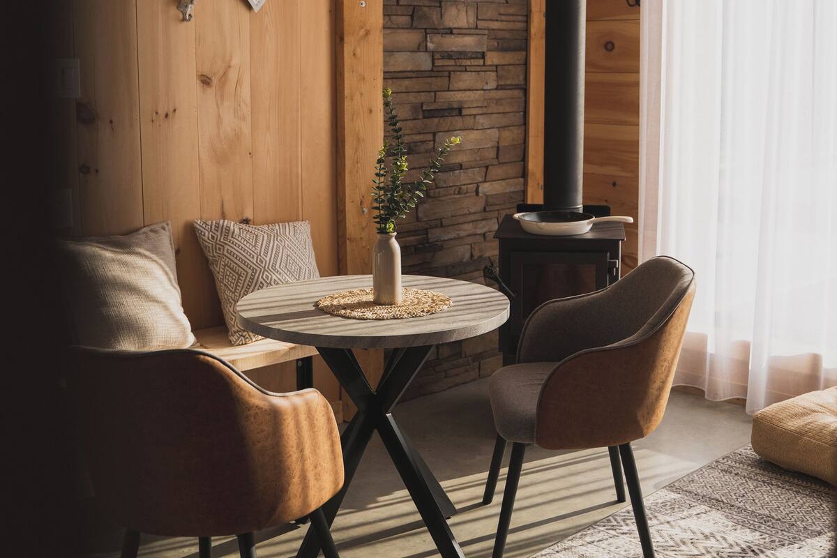Um quarto acolhedor com uma mesa redonda, duas cadeiras, um sofá, uma parede de pedra, uma parede de madeira e uma chaminé preta.