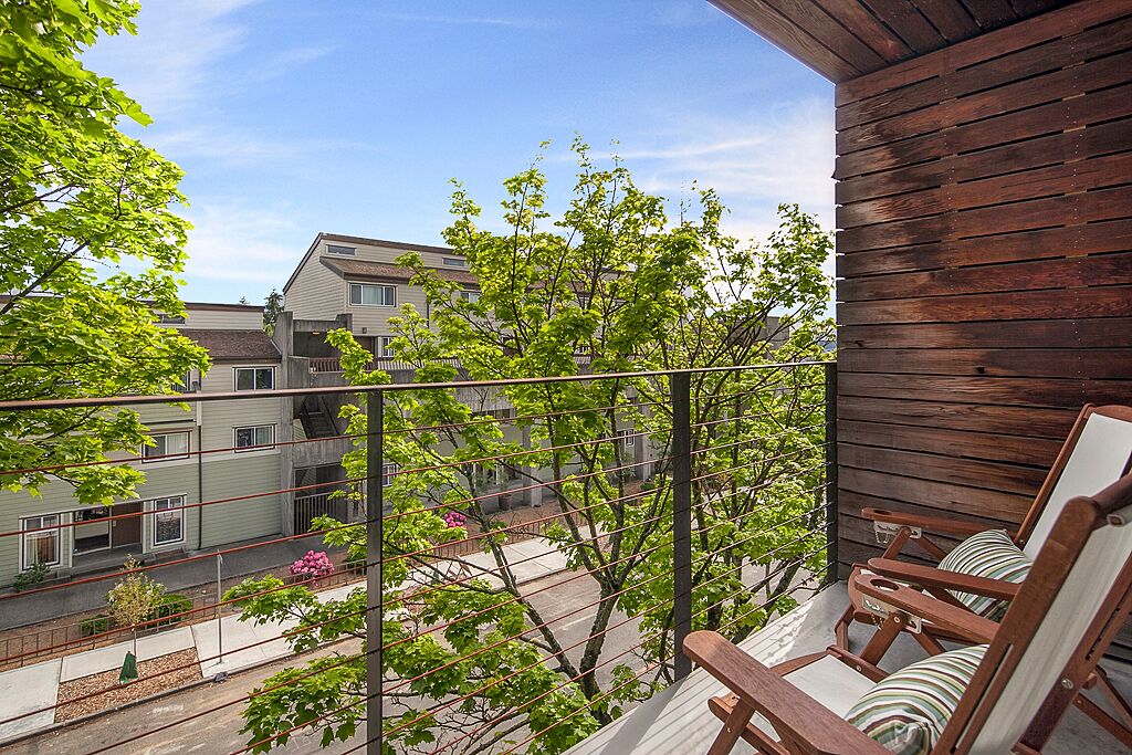 A balcony with wooden railings, a view of neighboring buildings, and two wooden chairs with cushions.