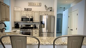 A modern kitchen with granite countertops, stainless steel appliances, and a farmhouse sink. There are two barstools and a sign that reads "FAMILY" above the cabinets. - Wildflower Ranch Backyard Oasis with Pool & Spa (Goodyear)