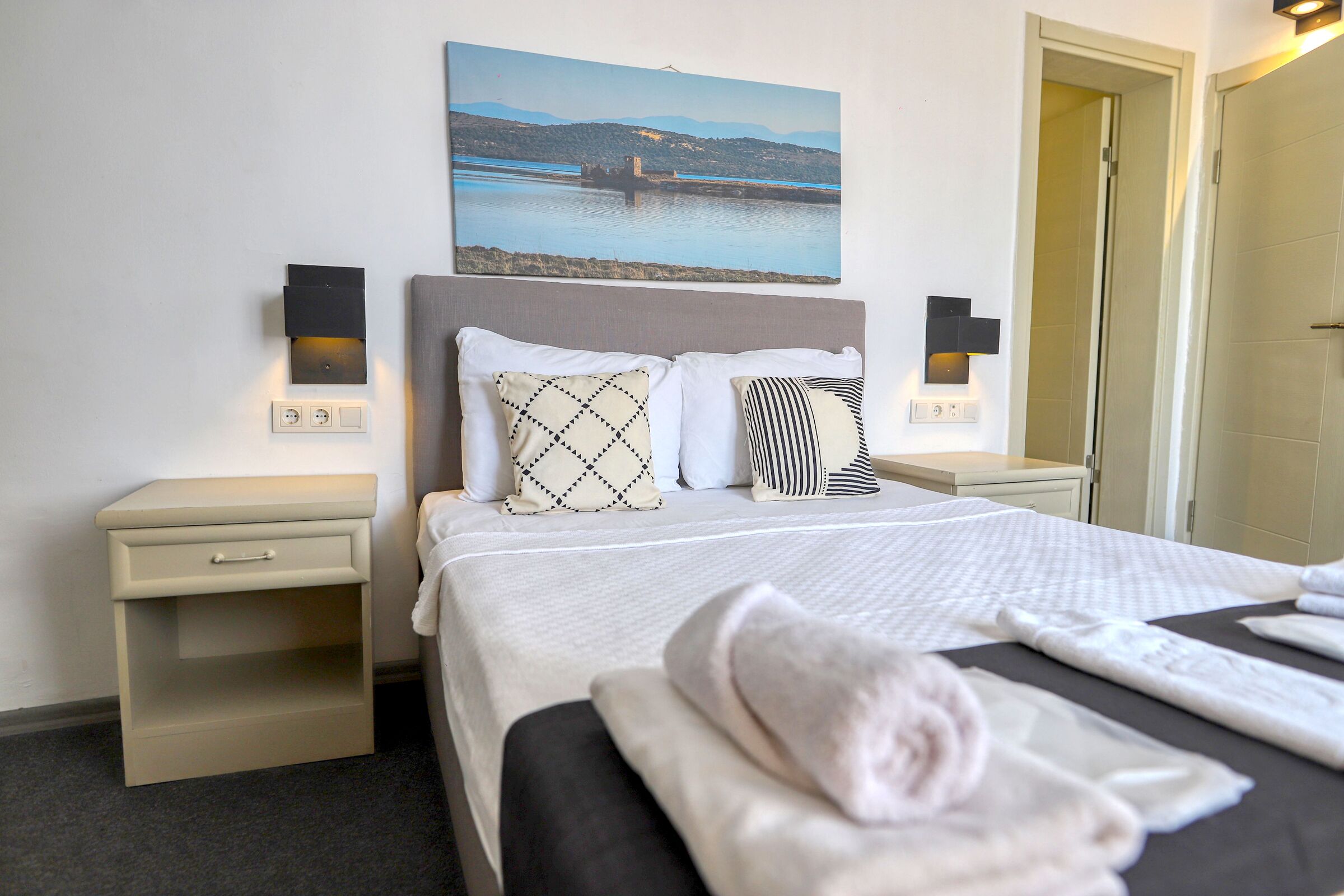 A neatly made bed with a white and black patterned pillow, a folded towel, and a painting of a landscape on the wall.