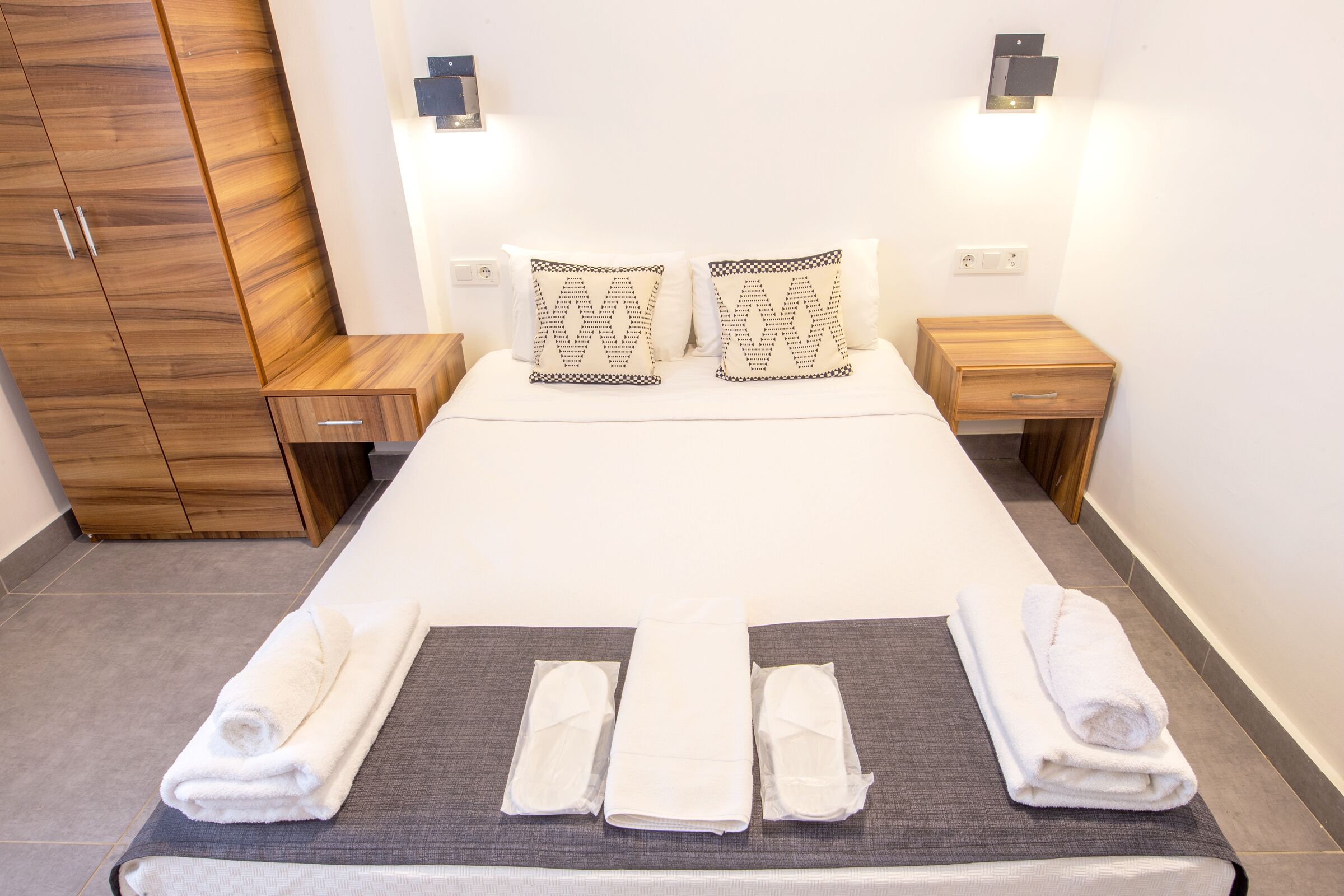 A neatly made bed with white linens, two decorative pillows, and a set of towels arranged on a gray mat.