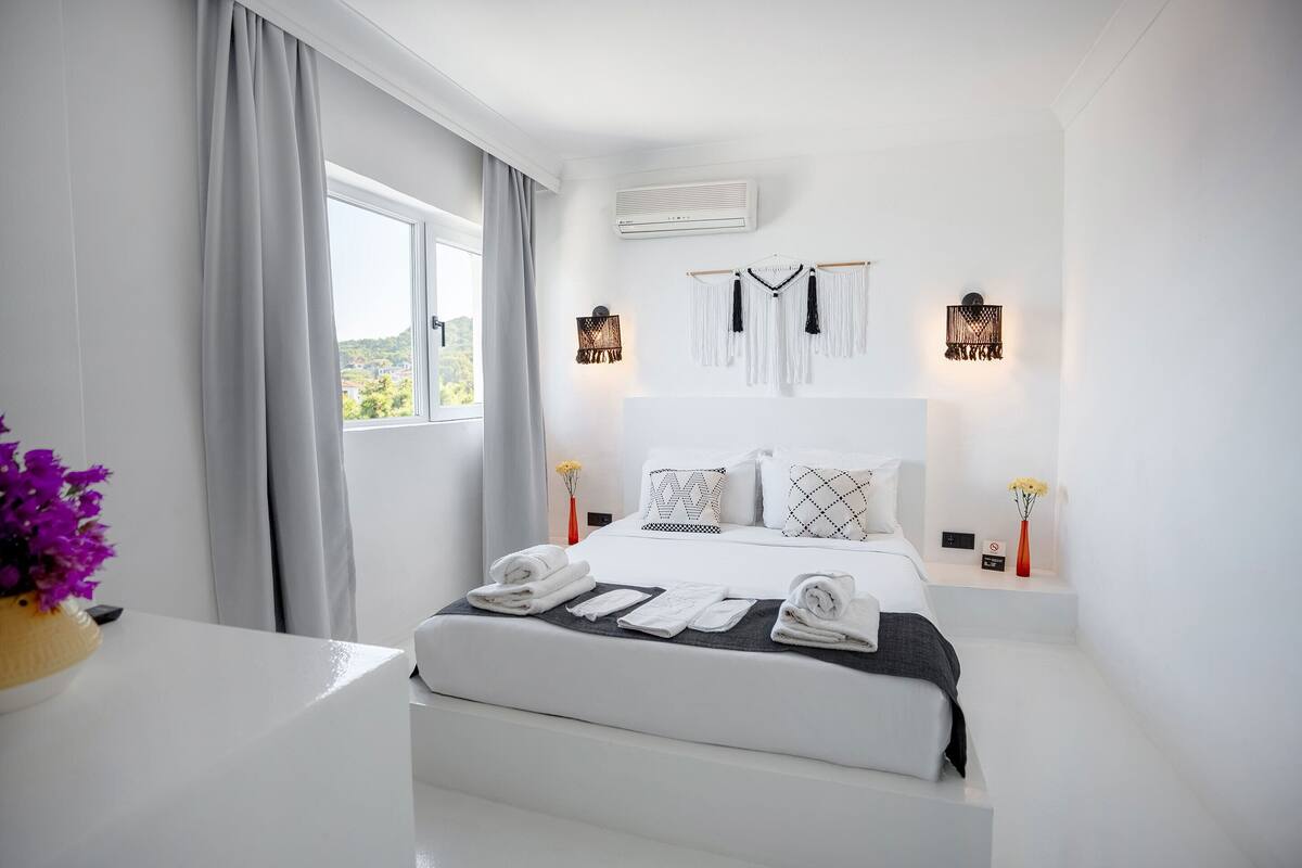 Une chambre d’hôtel moderne avec un lit bien fait, une grande fenêtre avec des rideaux, un climatiseur fixé au mur et deux appliques murales.