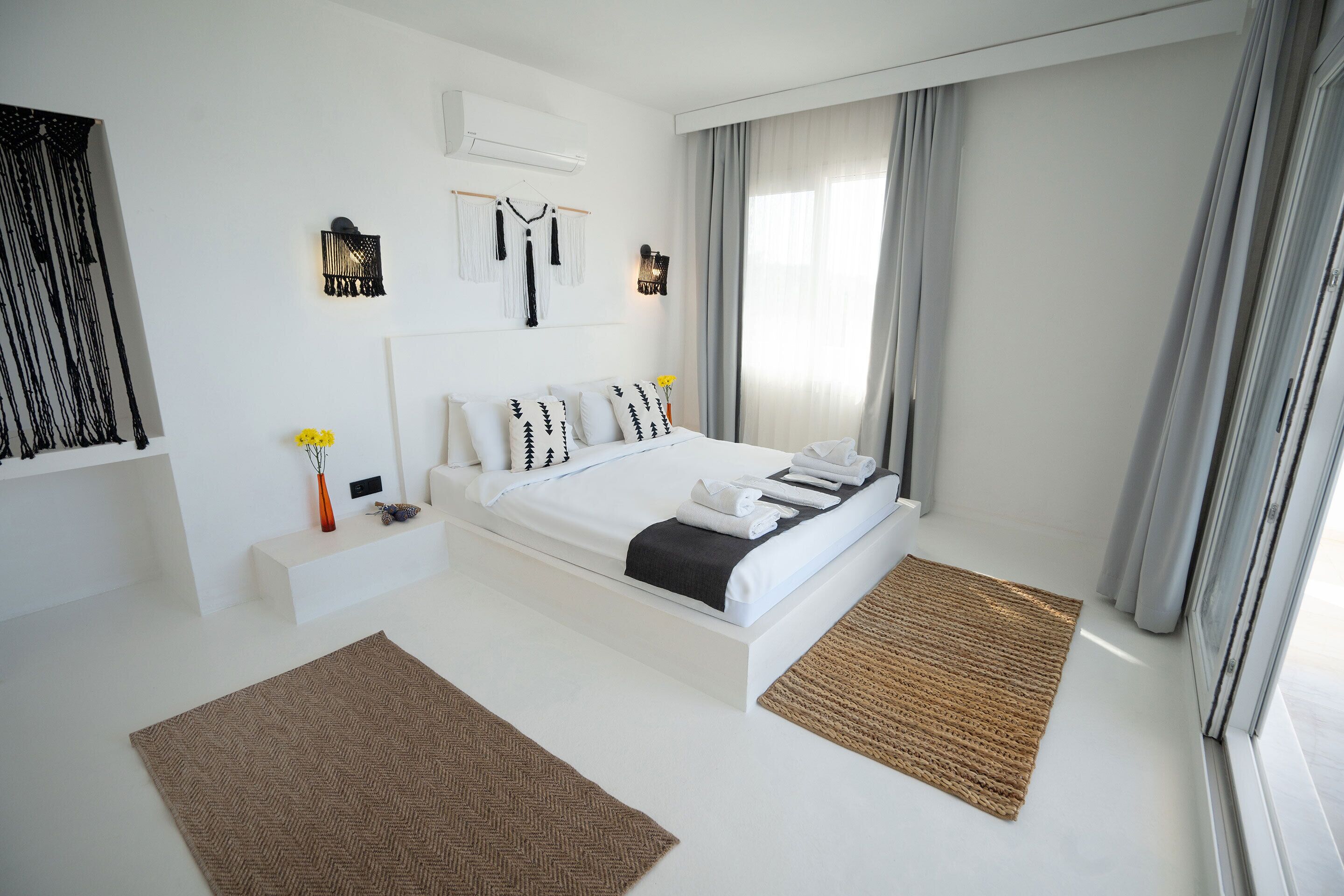 A modern hotel room with a large bed, a small bedside table, a wall-mounted air conditioner, and a window with curtains.