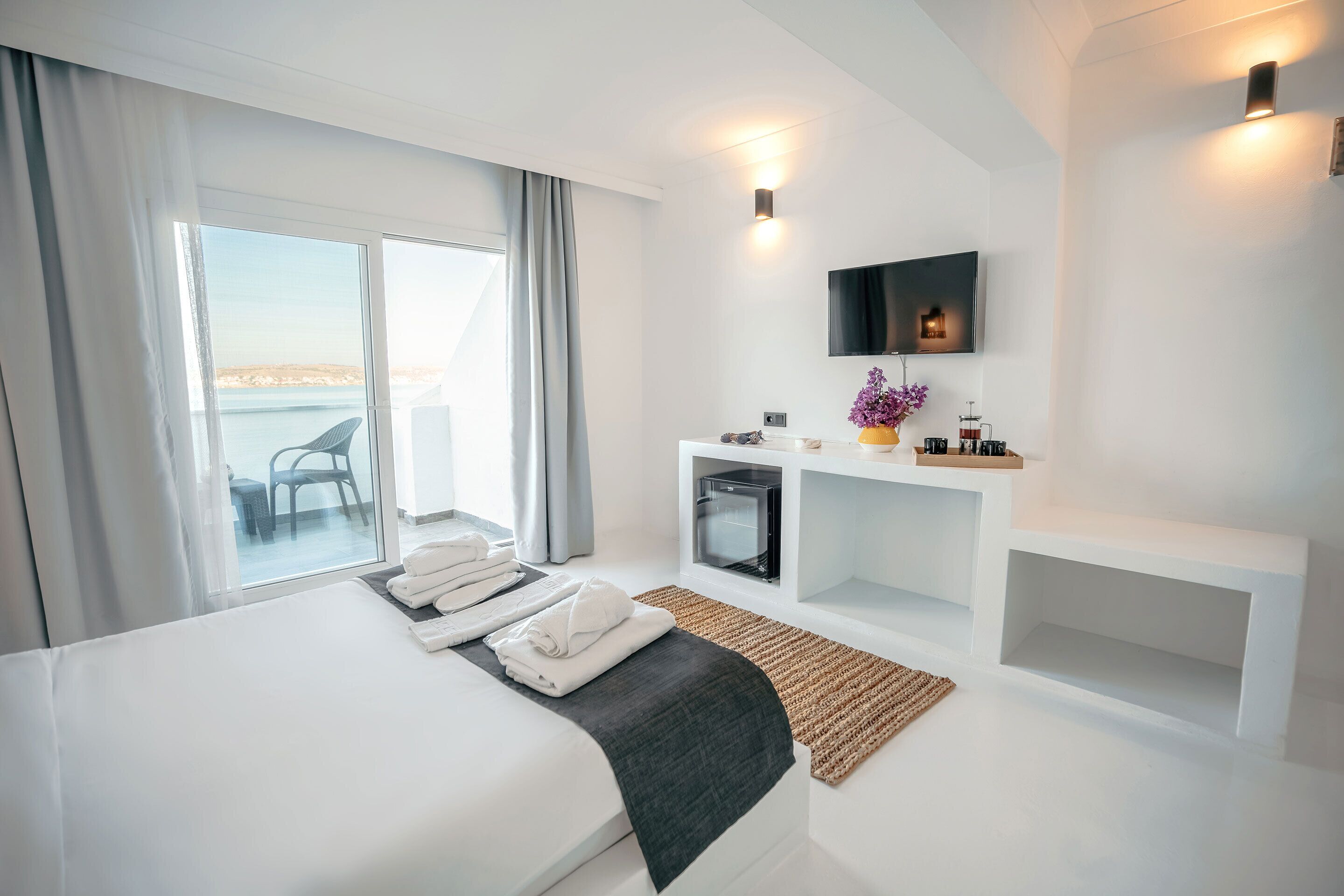 Une chambre d’hôtel moderne avec un grand lit, un téléviseur fixé au mur, un petit bureau agrémenté d’un vase de fleurs et une vue sur la mer par la fenêtre.